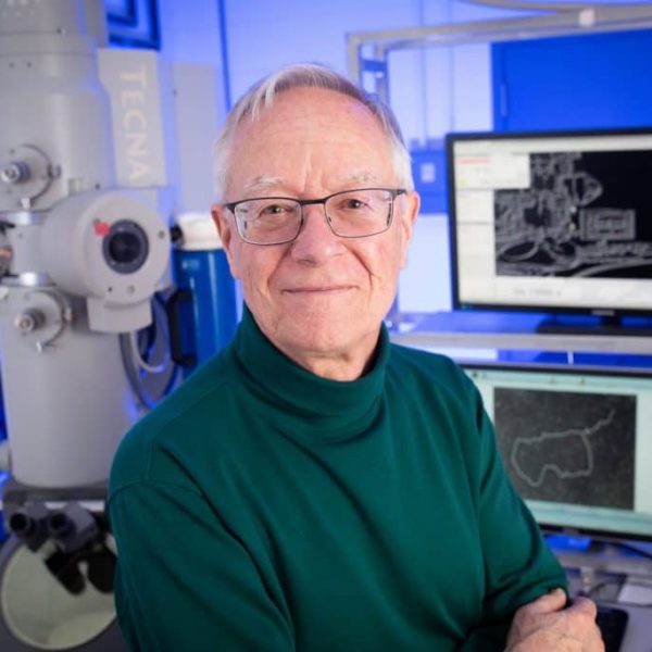A man with short grey hair wearing glasses and a dark green turtleneck stands in front of electron microscope imaging equipment.