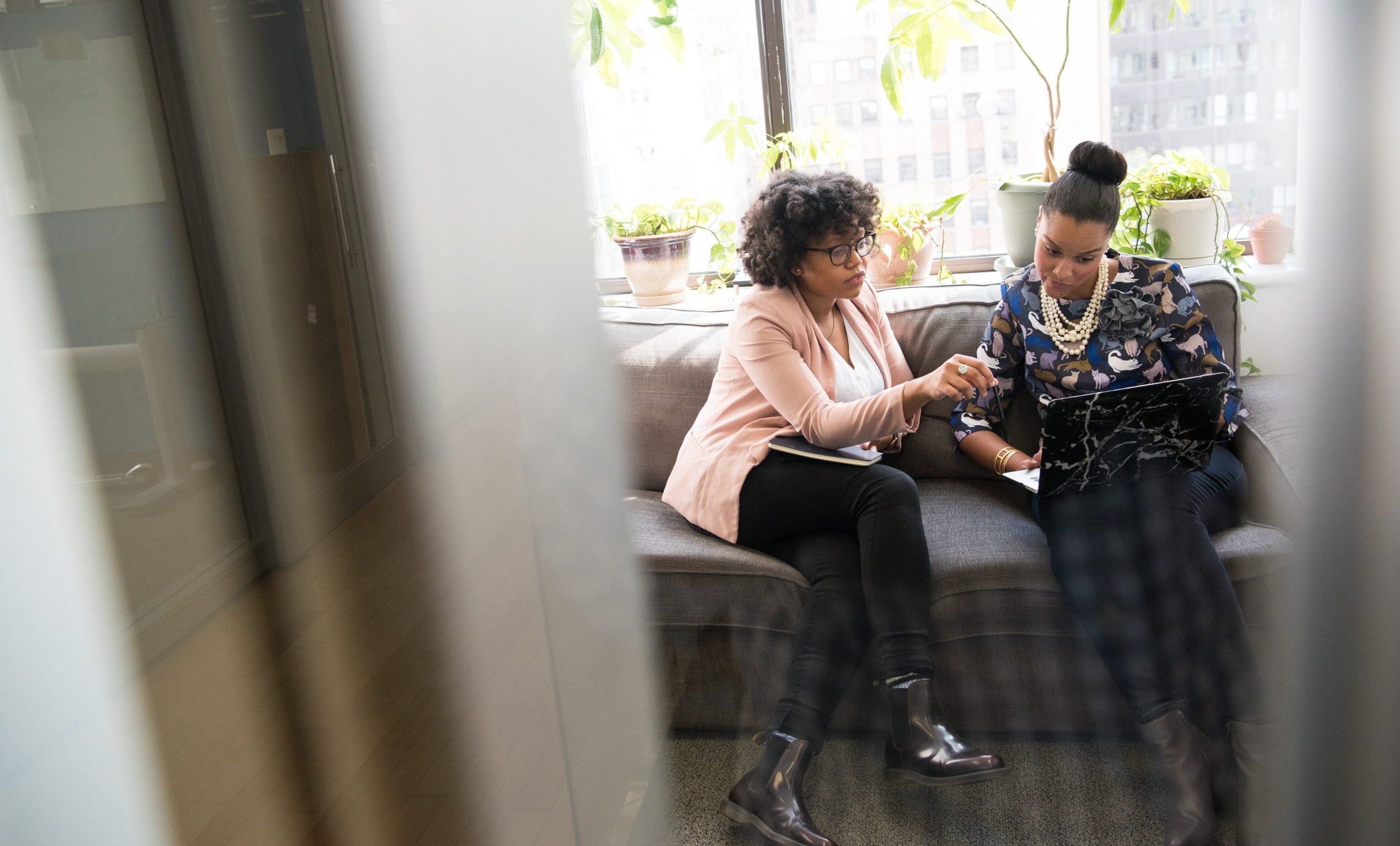Two black women in business casual clothing sit on a sofa in an office setting. They are looking at a laptop together.