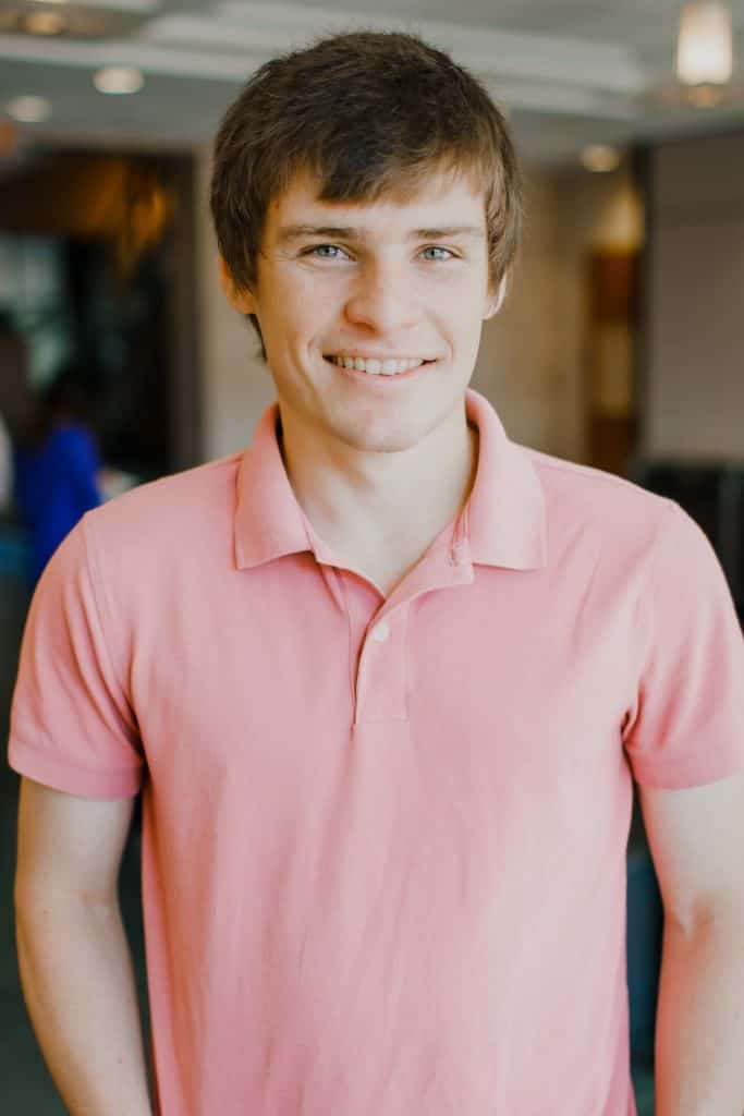 A smiling man with short light brown hair. He is wearing a light pink polo shirt.
