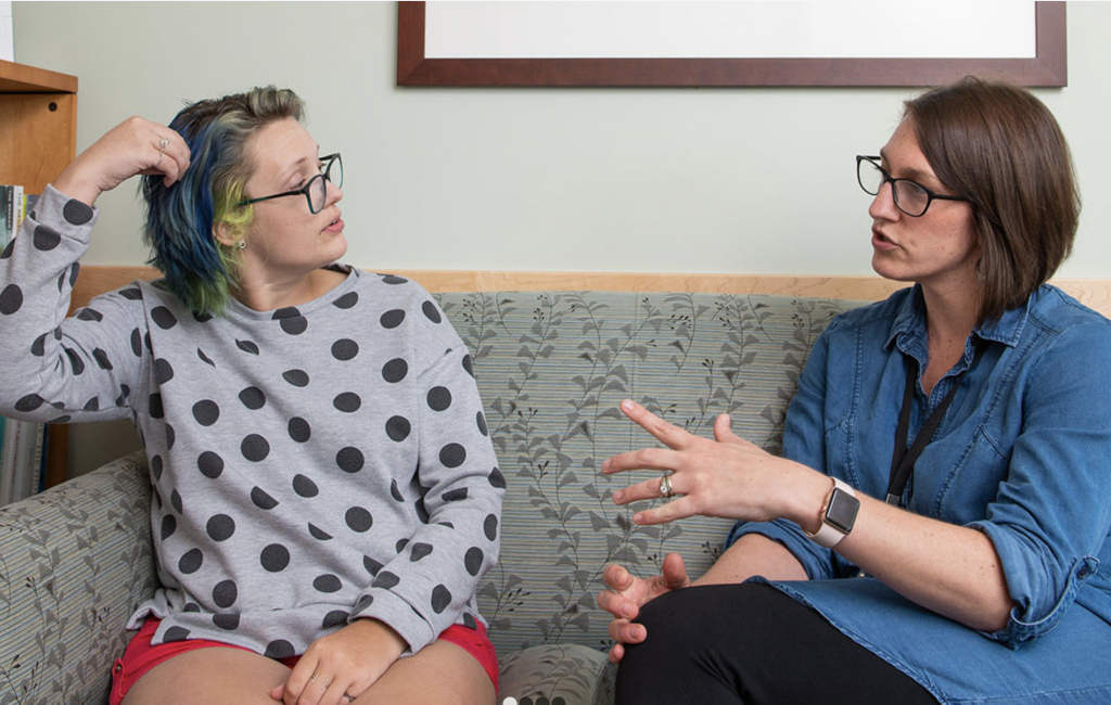 Young adult cancer patient sits and is talking with Program Coordinator Lauren Lux on a sofa.