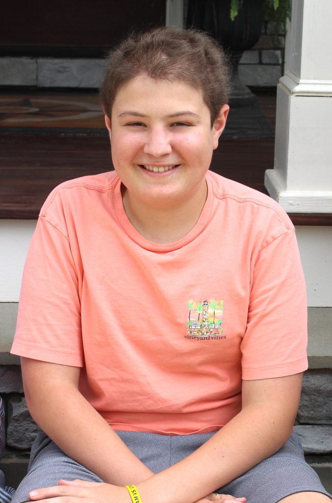 A smiling teen boy sits on the steps of his home's front porch, wearing a coral-colored t-shirt and grey shorts.