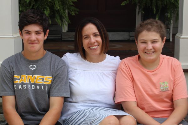 Three family members sit next to each other on the steps of their porch. The two brothers and their mom all have brown hair and are smiling.