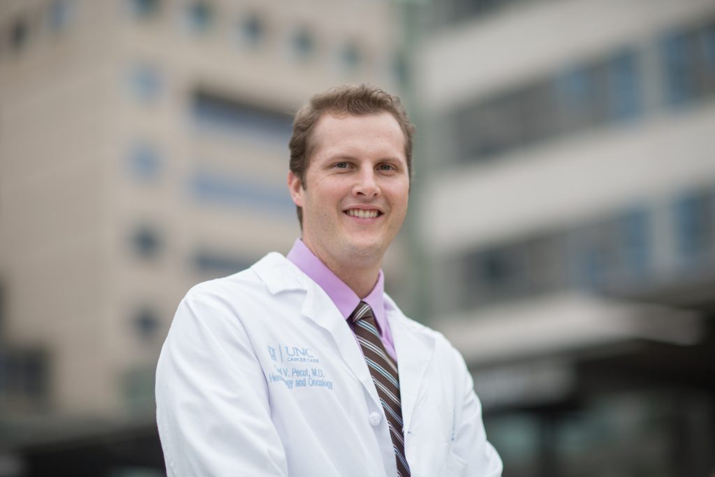 A smiling man with short blonde hair wearing a pink buttoned shirt with a striped tie and a white physicians coat. He is outside with the exterior of a healthcare building in the background.