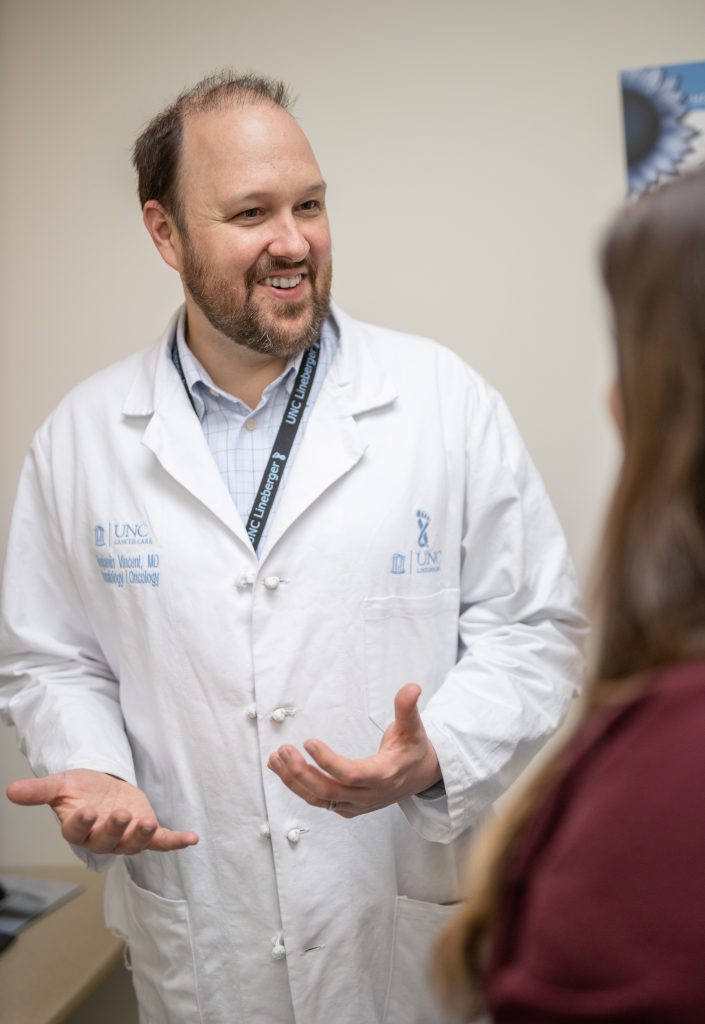 Ben Vincent wearing a doctor's white coat talking with a patient in an exam room