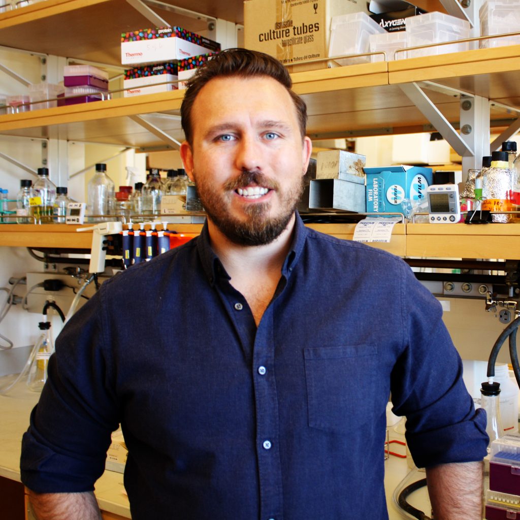 A smiling man with brown hair and a beard wearing a navy blue buttoned shirt standing in front of a lab bench with shelves full of research supplies.