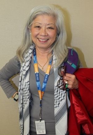 A smiling woman with gray shoulder-length hair wearing a gray shirt and patterned scarf.