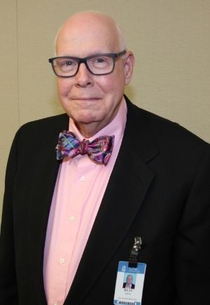 A man wearing glasses, a light pink buttoned shirt and pink bow tie with a black jacket.