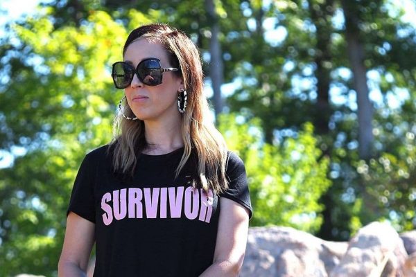 A brunette woman stands outside wearing oversized sunglasses, hoop earrings, a black t-shirt with the word "survivor" in light pink text. She is looking to the side, away from the camera.