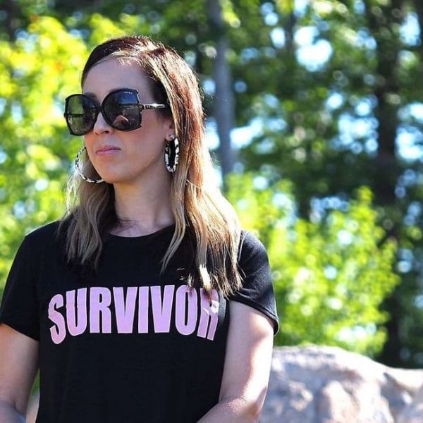 A brunette woman stands outside wearing oversized sunglasses, hoop earrings, a black t-shirt with the word "survivor" in light pink text. She is looking to the side, away from the camera.