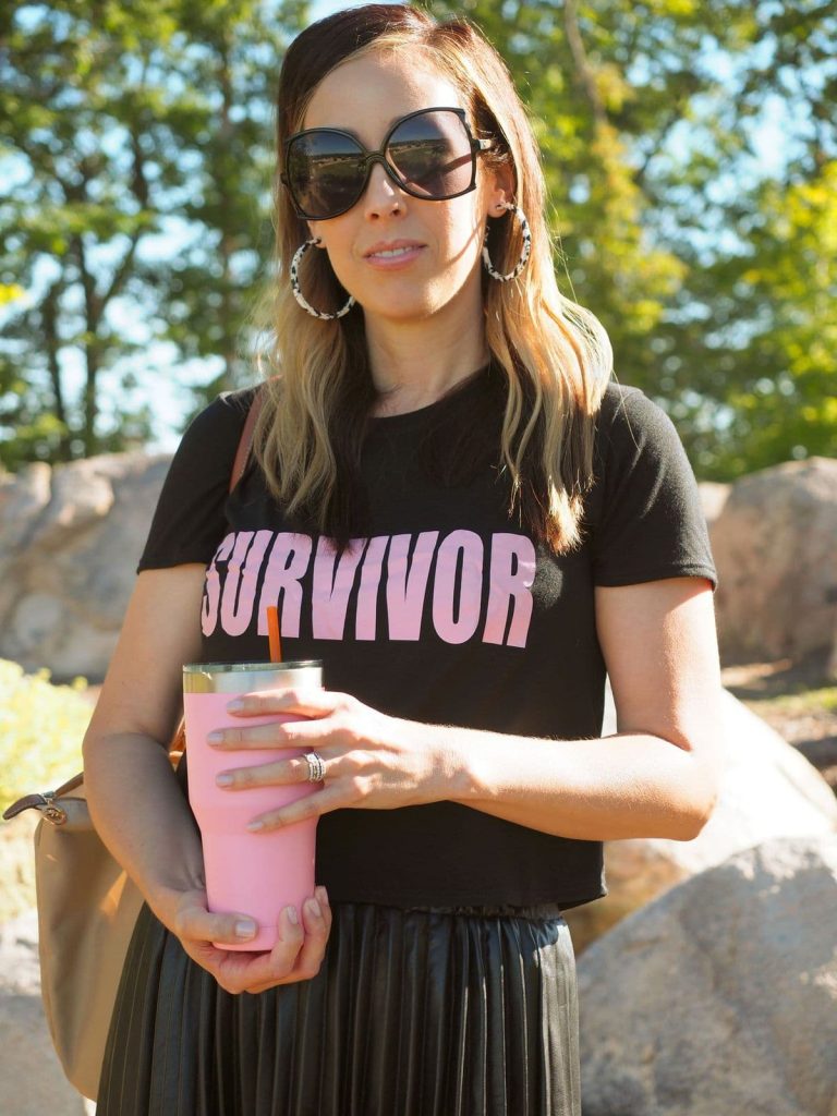 A brunette woman stands outside wearing oversized sunglasses, hoop earrings, a black t-shirt with the word "survivor" in light pink text. She is holding a light pink beverage tumbler.