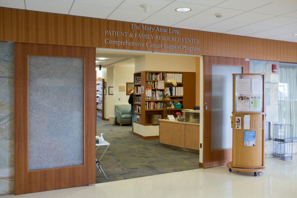 The entrance of the Patient and Family Resource Center in the N.C. Cancer Hospital. A wood panel wall with double doors propped open, looking inside the PFRC and showing bookshelves with pamphlets and the front desk.
