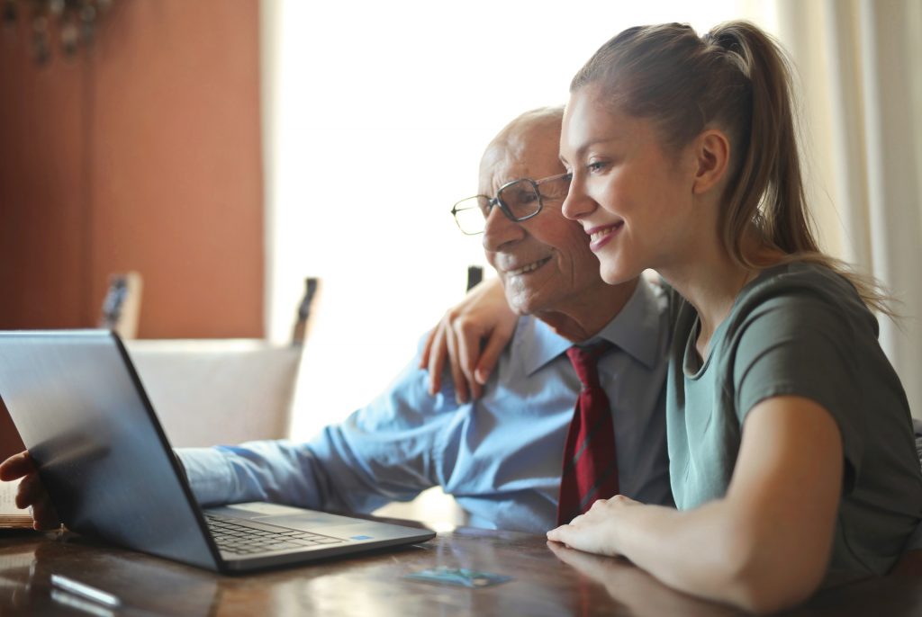 A man sits at a laptop with his caregiver. She has her arm around him and they are both smiling, looking at the laptop screen.
