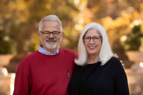 A smiling couple stands outside with an autumn background. Both people have white hair and wear glasses. They are wearing sweaters and the man has his around affectionately wrapped around the woman's shoulder.