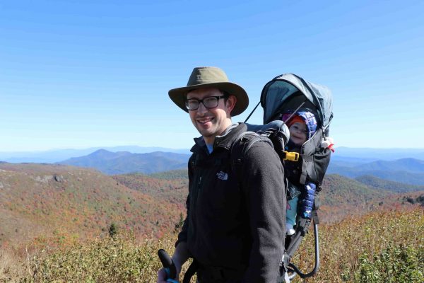 A smiling man wearing a rimmed hat stands on a mountain overlook. He is wearing glasses, a long-sleeved shirt, and is carrying a baby in a child-carrying hiking pack on his back.