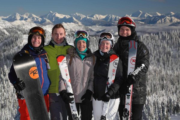 A group of smiling people stand outside with snow-covered mountains behind them. They are wearing winter sport outdoor wear and holding skiis and snowboards.