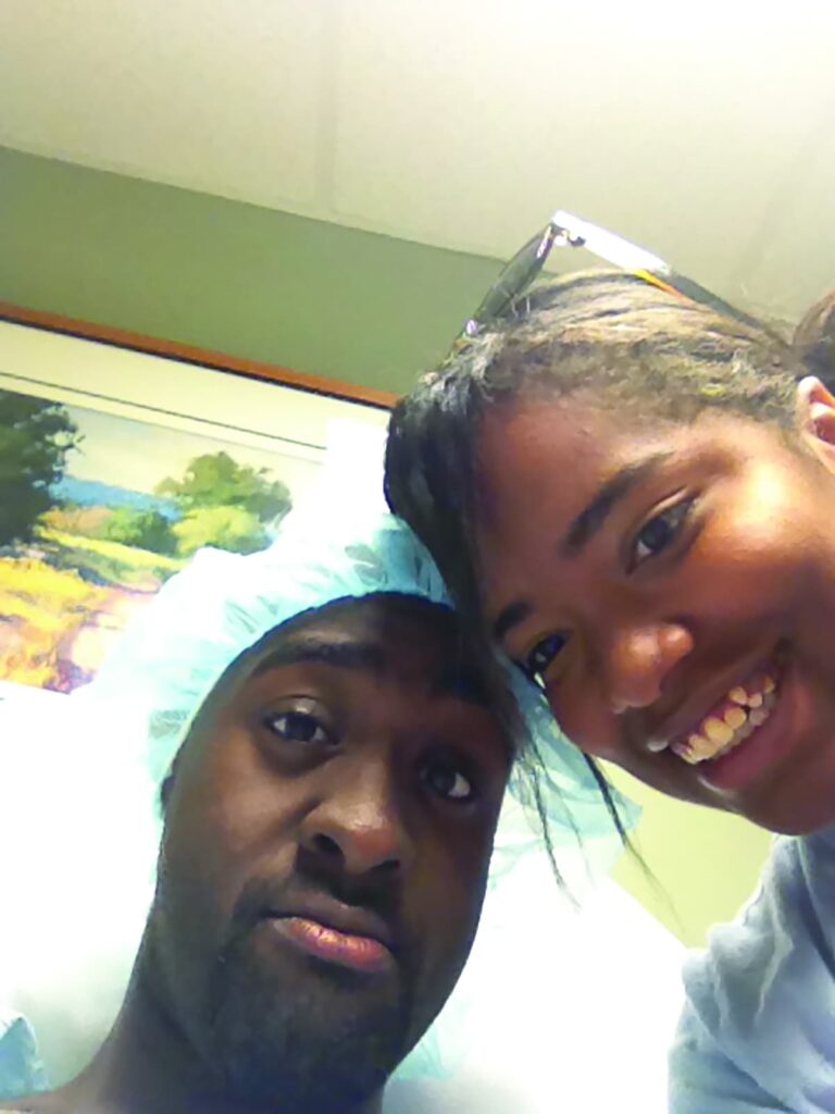 A selfie of two people smiling in a hospital room. One person is wearing patient pre-surgery garments.