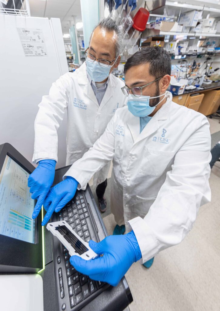 Two researchers wearing white coats, masks and gloves review data points on a computer screen.