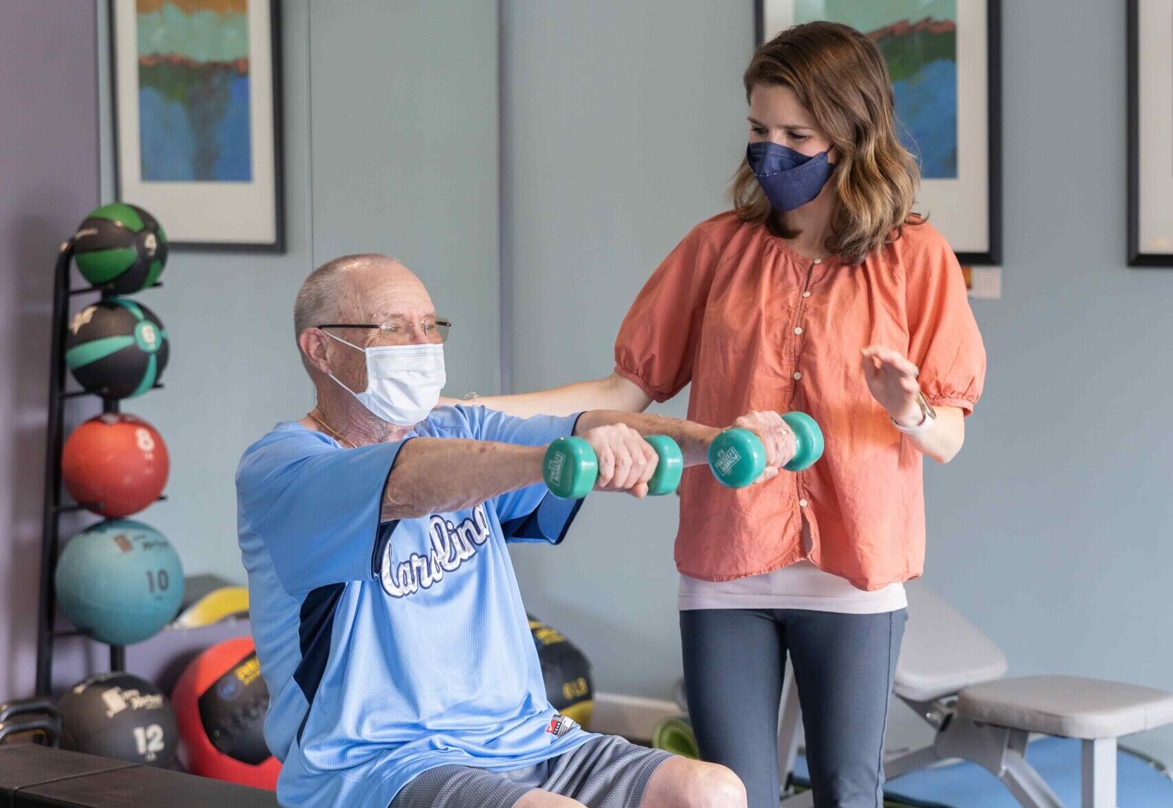 A patient works with a trainer to lift hand weights