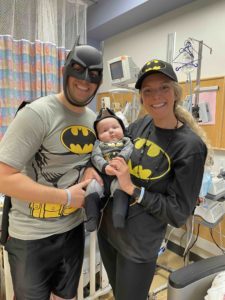 Parents with an infant, all dressed in batman costumes inside a hospital room.