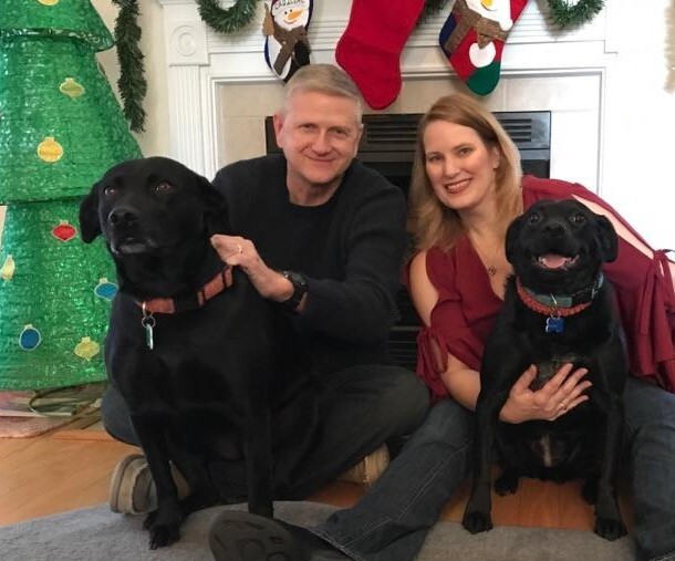 A man and woman with two black dogs in front of Christmas decorations
