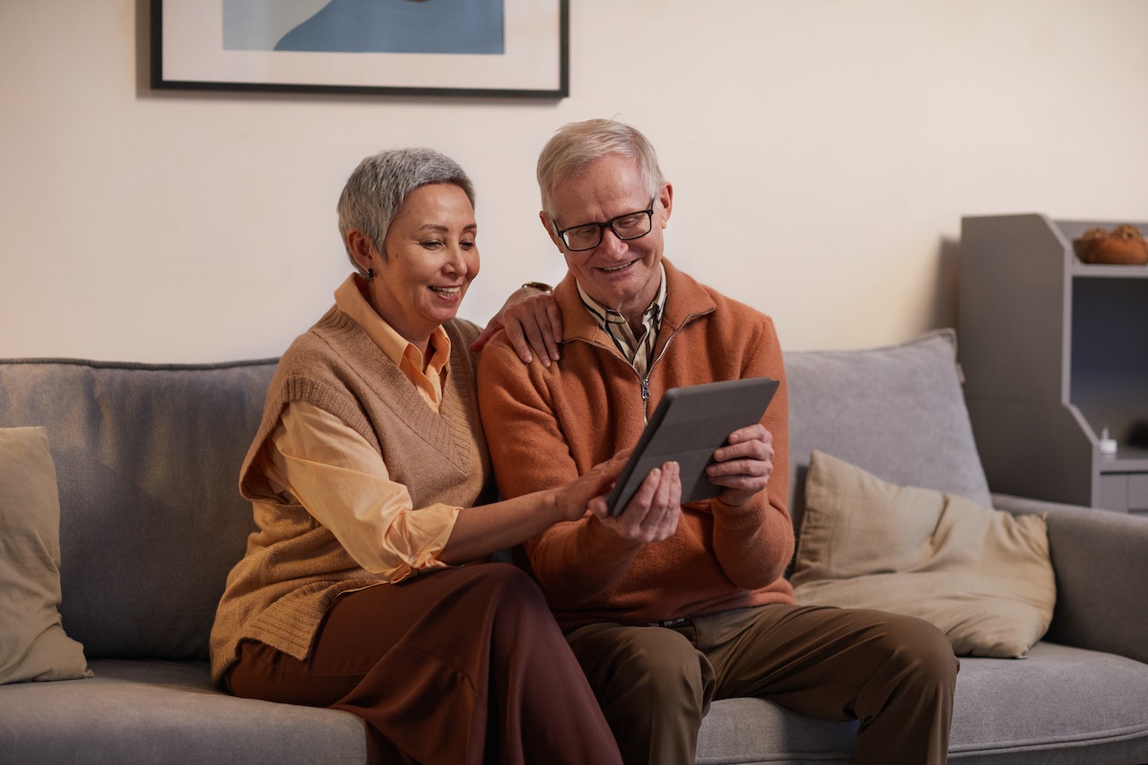 A couple sits on a sofa, smiling and holding a tablet.