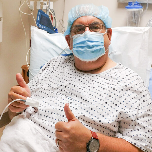 John Bell in his hospital room, wearing a patient gown and gesturing with two thumbs up.