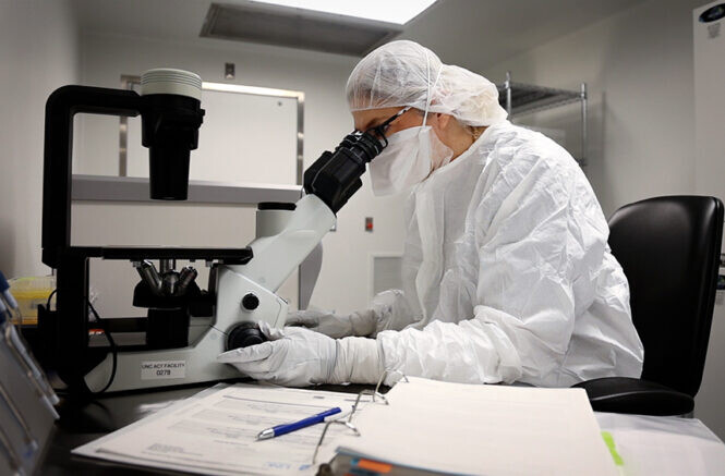 A person wearing PPE looking into a microscope.