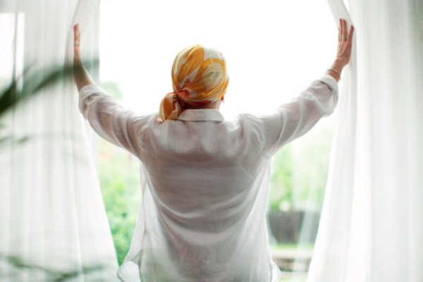 A person wearing a headscarf opens window curtains, looking out on a sunny day.